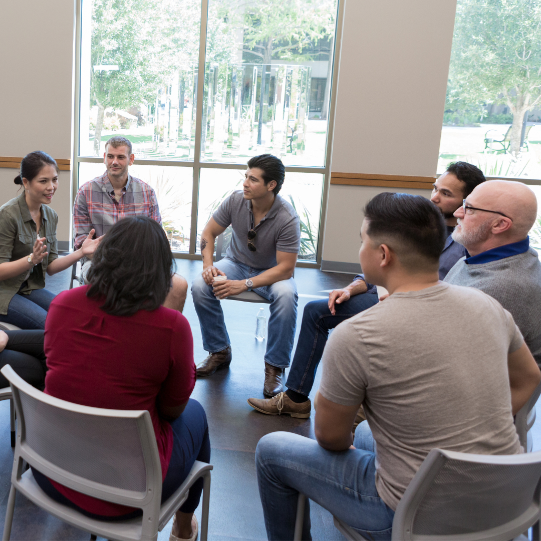 A group of people sitting in a circle, having a discussion in a well-lit room with large windows providing a natural view.
